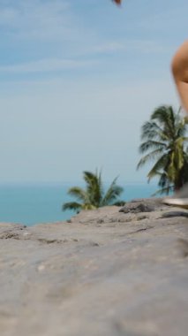 Dikey video. Kayalık bir bakış açısıyla yürüyen, arka planında parlak gün ışığında palmiye ağaçları ve okyanus olan, alçak açılı, çakışan taş Koh Samui konsepti olan kadın.