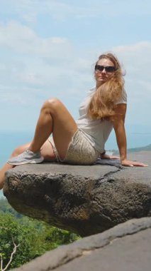 Dikey video. Üst üste binen taşın üzerinde oturan genç kadın geniş okyanus manzaralı ağaçlarla çevrili kenar açılı geniş açılı Samui Doğa Macerası