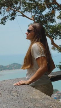 Dikey video. Güneş gözlüğü takan kadın Tahta parmaklıkların yanında duruyor Geniş Okyanus ve Tepeler 'in tadını çıkarıyor Üst Kaya Samui' de Yan Açıdan Panorama, Açık Hava Macera Konsepti