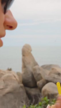 Dikey video. Samui Adası 'ndaki Kayalıklarda oturan Renkli Kaşıklı Hindistancevizi Dondurması Tatma Kadın Samui Adası' ndaki Hin Ta Hin Yai, Tropikal Tatlı Şeker Konsepti