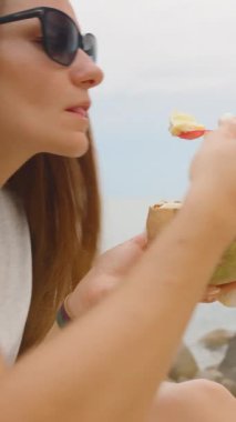 Dikey video. Güneş Gözlüklü Kadın Denize Yakın Oturan Çiçeklerle Hindistan cevizli dondurma ve Samui Adası 'nda Iconic Hin Ta Hin Yai Kayalıkları, Kenar Açı, Tropikal Muamele Konsepti