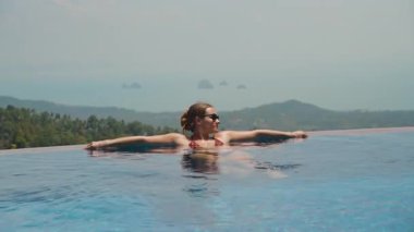 Yeşil Tepeler ve Okyanus Dinlenme Konsepti ile Sonsuzluk Havuzu 'nun Kıyısında Dinlenen Güneş Gözlüklü Kadın Görünümü