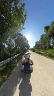 Dikey video. Erkek Sürücünün Arkasında Oturan Kadın Scooter 'la Orman Yolu' nda Güneş 'e doğru gidiyor. Arka Görüş Perspektifi, Tropikal Seyahat Konsepti