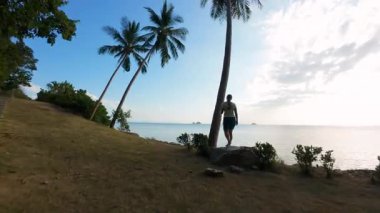 Palmiye ağacının yanında dikilen ve FPV İHA 'sı içinden geçip Horizon' a doğru uçarken okyanusu izleyen kadın, sinematik kaçış kavramı.