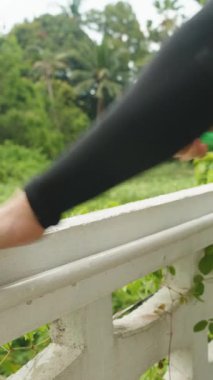 Dikey video. Açık Hava Balkonunu Temizleyen Adam Parlak Gök Altındaki Tropikal Bitkilerle çevrili Hortum Spreyle Korkuluk