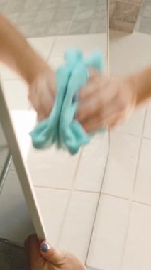 Dikey video. Banyoda aynanın altını mavi mikrofiber bezle silen kadın. Düşük açı, yüzey parlatıcı konsept.