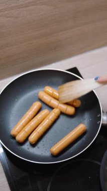 Dikey video. Küçük Mutfak 'taki Siyah Elektrikli Soba' da Kızartma Tavasında Tahta Spatula Karıştırma Sosisleri, Üst Manzara, Fast Food Konsepti