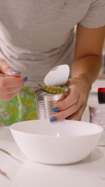 Dikey video. Mutfak tezgahında kaşıkla konserve bezelyeleri kepçeleyen kadın, yakın çekim açısı, ev yemeği kavramı.