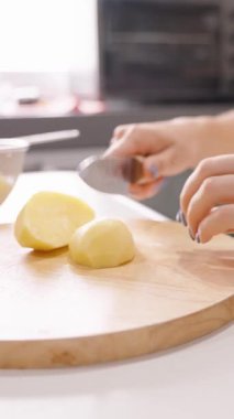 Dikey video. Kadın Soyulmuş Patatesleri Tahta Kesme Tahtasına Dilimliyor. Yanında Cam Kase, yan açı, Ev Yemeği Konsepti.