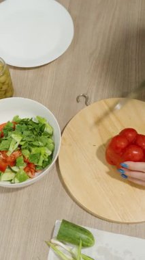 Dikey video. Çiçekli önlüklü bir kadının ahşap kesme tahtasındaki kırmızı dolma biberi dilimleyişini ve yanında bir kase taze sebze salatası, sağlıklı beslenme konsepti.