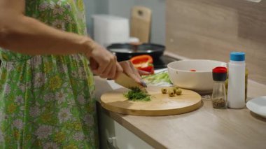 Yeşil önlüklü bir kadının mutfak tezgahında salata zeytin ve baharat kasesinin yanında taze dereotu doğraması, sağlıklı beslenme konsepti.