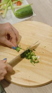 Dikey video. Ahşap bir kesme tahtasının üzerinde yeşil soğan doğrayan el pozu içinde salatalık, yeşillik ve salatalık içeren büyük bir kase salata, sağlıklı beslenme konsepti.