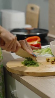 Dikey video. Yeşil önlüklü bir kadının mutfak tezgahında salata zeytin ve baharat kasesinin yanında taze dereotu doğraması, sağlıklı beslenme konsepti.