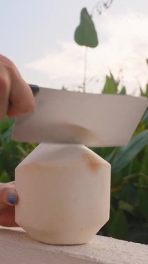 Dikey video. Çiçekli Kıyafetli Kadın Balkonda Büyük Bıçaklı Taze Hindistan Cevizi Kesiyor, Tropikal Yaşam Tarzı Konsepti