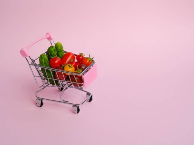 Pembe arka planda domates ve salatalık dolu pembe market arabası.
