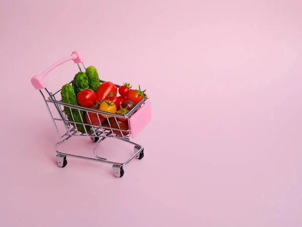 Pink grocery cart full of tomatoes and cucumbers on pink background