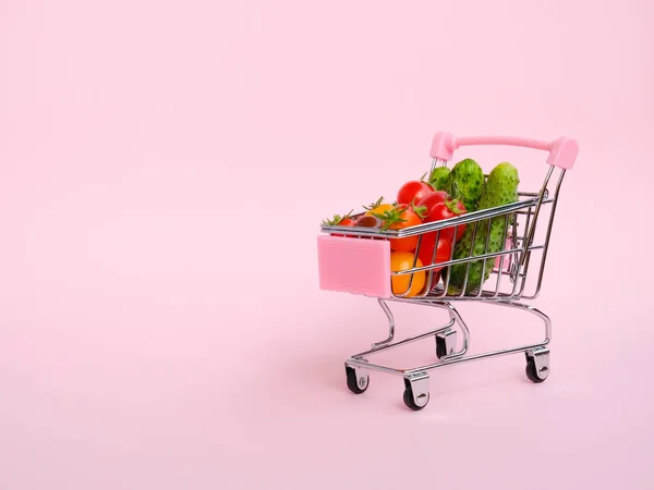 Pink grocery cart full of tomatoes and cucumbers on pink background