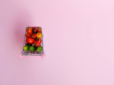 Pembe arka planda domates ve salatalık dolu pembe market arabası.