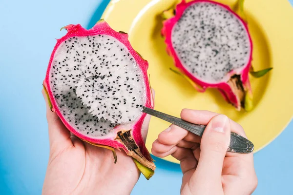 Pitaya is the fruit of the dragon. The girl is eating a tropical fruit with a spoon. Sweet pitaya on a blue background. Yellow plate and cut fruit nutrition. Dragon fruit close-up