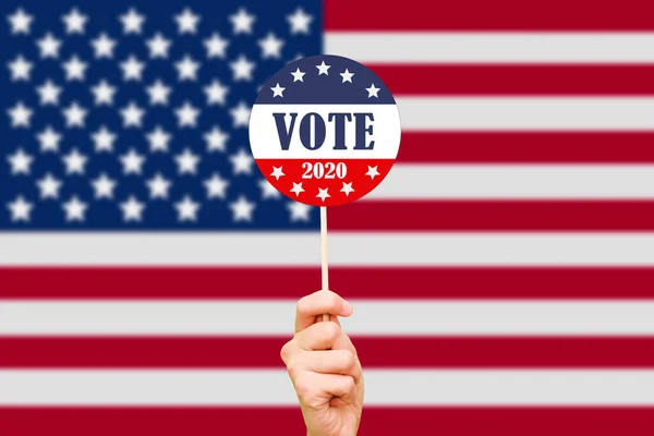 Mano y botón para votar en las elecciones en el fondo de la bandera estadounidense. Las elecciones presidenciales de Estados Unidos de 2020. Día de votación en los Estados Unidos de América — Foto de Stock