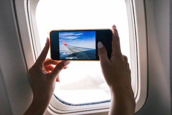 Yolcu uçakta fotoğraf çekiyor. Kız, uçak penceresinden cep telefonu kamerasıyla fotoğraf çekiyor. Eller yakın çekim ve akıllı telefon ekranı. Seyahat ve teknoloji.