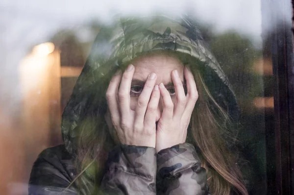 Beautiful woman with hooded jacket is worried and looking through the window and holding her head