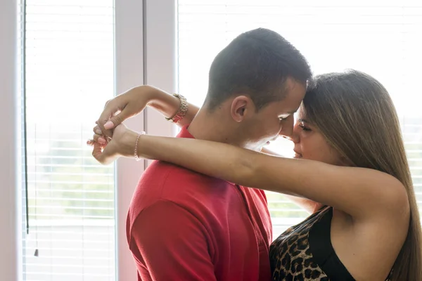 Young attractive couple is full of emotions, standing next to window, hugging and kissing