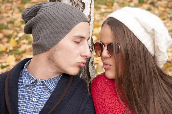 Young attractive casual dressed couple with woolen hats having a fun in public park beside the tree. Autumn time, fallen leaves.