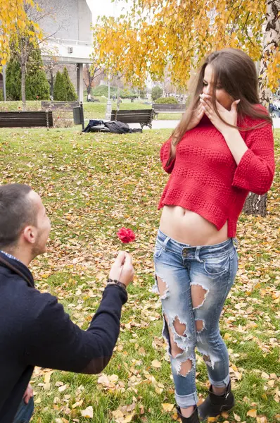 Young seductive woman in torn jeans is excited while her boyfriend is giving her a flower, kneeling on the ground covered with fallen leaves. Autumn time.