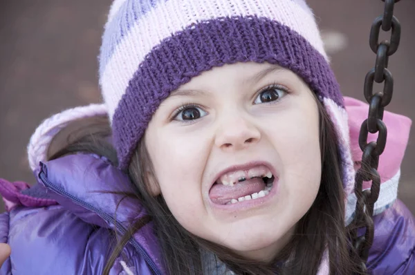 Sweet little girl in purple jacket and woolen purple hat making funny face on the swing