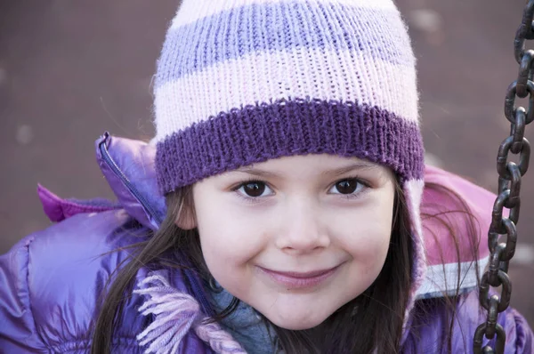 Sweet little girl in purple jacket and woolen purple hat having fun on the swing