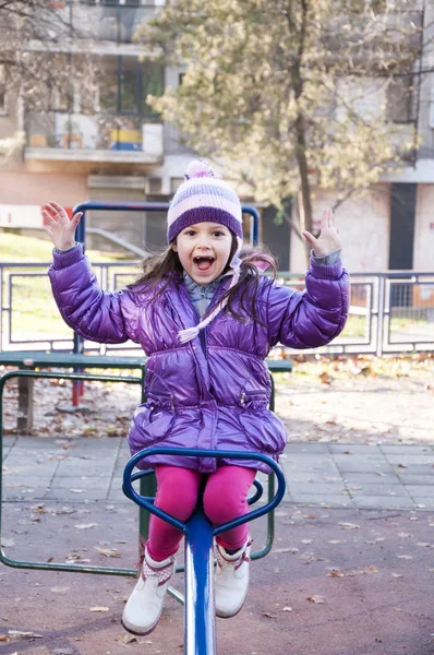 Sweet little girl in purple jacket and woolen purple hat having fun in the playground