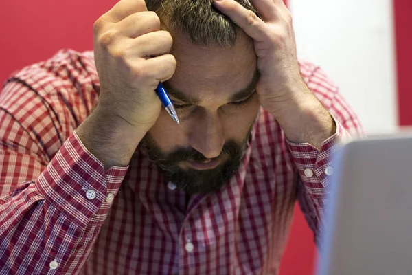 Portrait of disappointed bearded businessman in red and white shirt in the office holding his head.