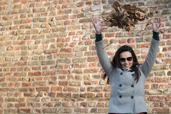 Attractive young woman near the brick wall throwing leaves