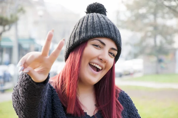 Portrait of seductive young redhead with woolen hat making a victory sign