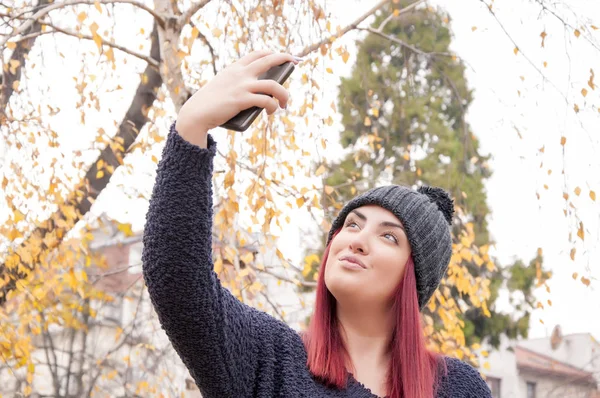 Seductive young redhead with woolen hat taking a selfie photo in the public park
