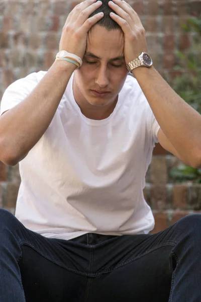 Outdoor portrait of sad young man covering his face with hands sitting on stairs. Selective focus on hands. Sadness, despair, tragedy concept