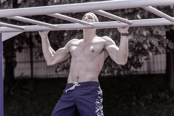 Young Muscular Man Doing Pull Ups in Public Park on Horizontal Bar