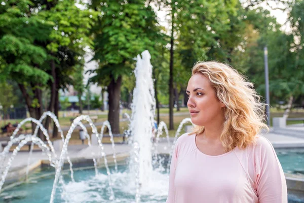 Modern young woman smiling by fountain in the public park on a bright sunny day.
