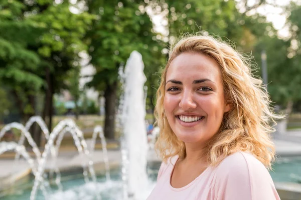 Modern young woman smiling by fountain in the public park on a bright sunny day.