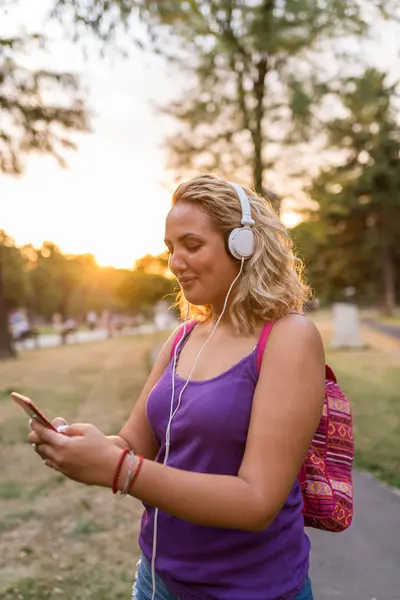 Woman with violet shirt and backpack using her smartphone outdoors during sunset.