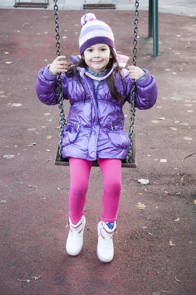 Sweet little girl in purple jacket and woolen purple hat having fun in the playground