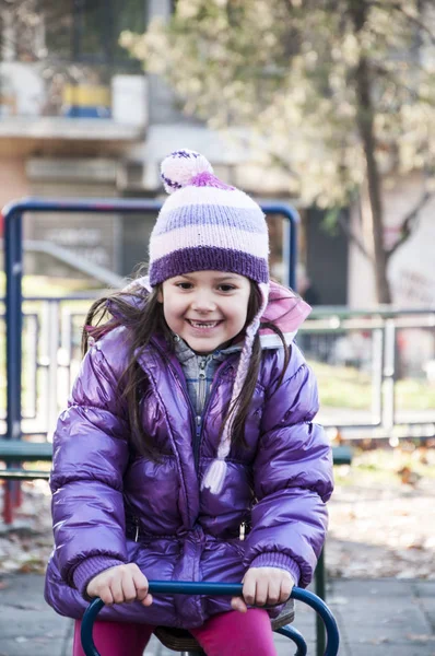 Sweet little girl in purple jacket and woolen purple hat having fun in the playground