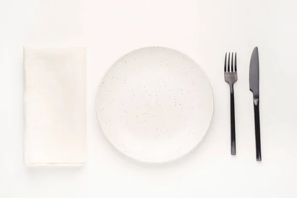 Set aus schwarzem Besteckmesser und Gabel, weißem Teller und Serviette auf weißem Tisch. — Stockfoto