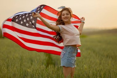 Pembe tişörtlü, üzerinde Amerikan bayrağı olan kot şortlu, gün batımında yeşil buğday tarlasında mutlu, güzel beyaz bir genç kız. Bağımsızlık Günü