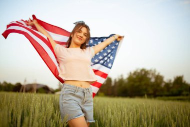 Pembe tişörtlü, üzerinde Amerikan bayrağı olan kot şortlu, gün batımında yeşil buğday tarlasında mutlu, güzel beyaz bir genç kız. Bağımsızlık Günü