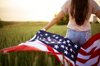 Pembe bluzlu, beyaz, genç, güzel, güzel bir kız. Üzerinde Amerikan bayrağı olan kot pantolonlar. Gün batımı manzaralı yeşil buğday tarlasında. Bağımsızlık Günü