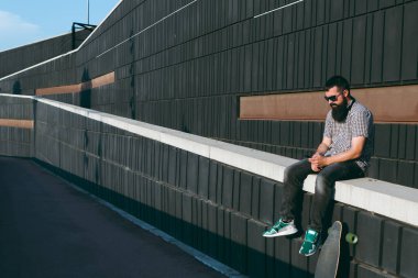 Sakal, kulaklık ve sokakta bir duvara oturan bir ekose gömlek ile genç patenci bir cep telefonu kullanıyor.