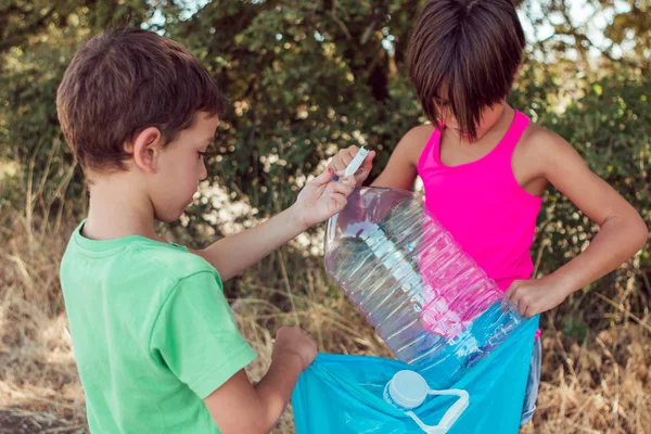Dos niños pequeños recogiendo una botella mientras sostienen una basura plástica en el bosque. Cuidar el concepto de medio ambiente — Foto de Stock