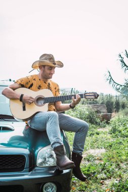 Açık havada gitar çalarken arazi aracının tepesinde oturan kovboy şapkalı genç bir çocuk.